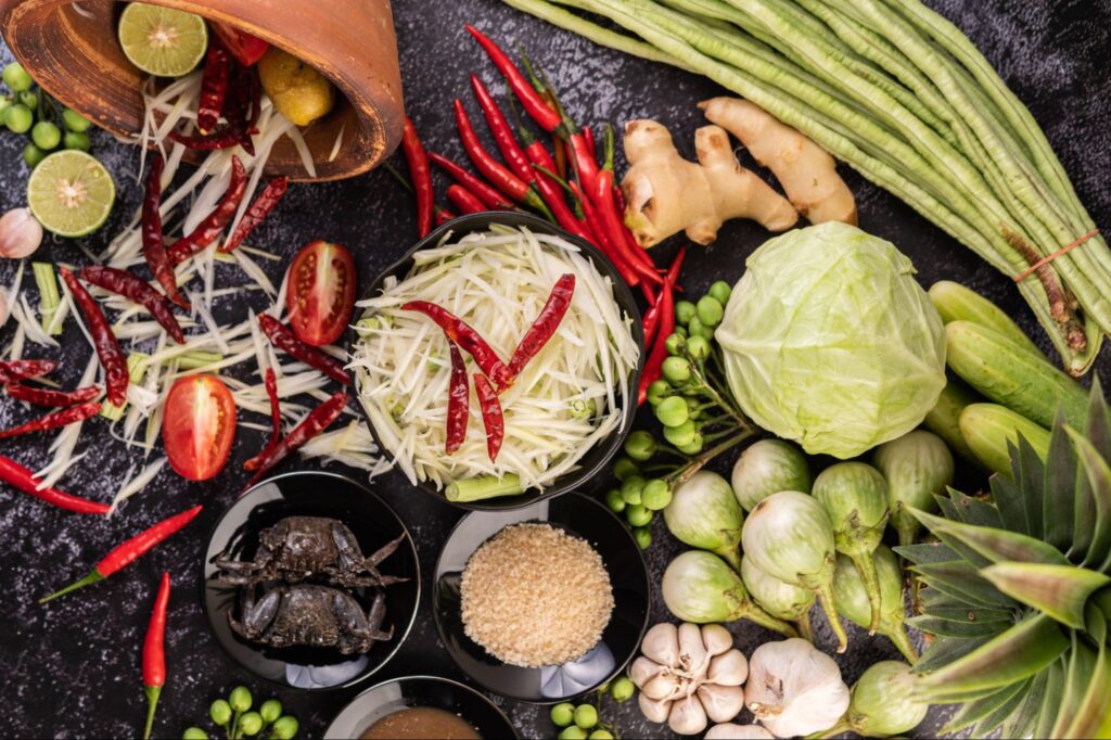 Flat lay of fresh Thai ingredients including shredded green papaya, red chillies, limes, ginger, long beans, cabbage, garlic, palm sugar, tomatoes, small crabs, and local vegetables arranged on a dark stone surface