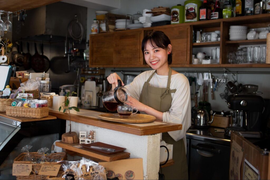 Smiling café barista in a neutral apron pouring freshly brewed coffee into a glass cup at a wooden counter, surrounded by shelves of cups, brewing tools, and warm café interiors.
