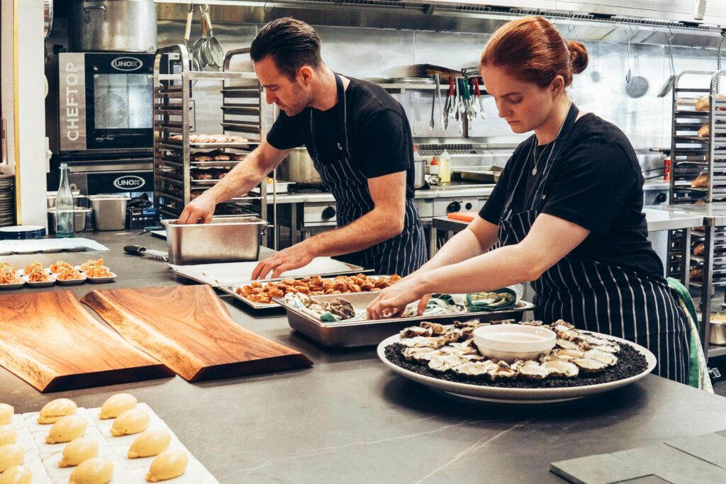 Two chefs in aprons carefully preparing plated dishes and fresh oysters on a stainless steel counter inside a busy commercial kitchen, with trays of food and cooking equipment in the background
